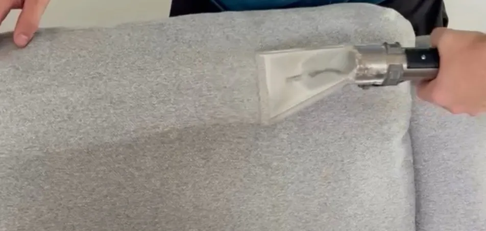 Upholstery tool cleaning a grey fabric couch cushion, leaving a lighter clean stripe
